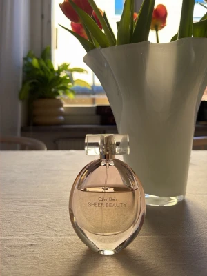 Calvin Klein Sheer Beauty parfym - En elegant parfym från Calvin Klein, Sheer Beauty, i en oval glasflaska med ljusrosa ton och transparent lock. Flaskan har en stilren och modern design som känns både fräsch och feminin.