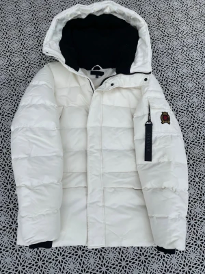 Vit pufferjacka från SikSilk - Säljer en snygg vit pufferjacka från SikSilk med huva och svart innerfoder. Jackan har en patch med broderad logga på ärmen och en dragkedjedetalj med SikSilk-tryck. Perfekt för kalla vinterdagar och riktigt trendig look. Jätte bra för kallar väder använd 4-5 gånger ett litet märke på höger arm går bort med lite tvätt hör av vid frågor 