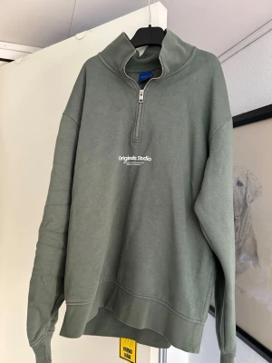 Grön halfzip hoodie från Jack & Jones - Snygg grön hoodie från Jack & Jones Originals med halv dragkedja och hög krage. Tillverkad i mjuk bomullsblandning och har trycket 'Originals Studio' på bröstet. Perfekt för en avslappnad och trendig stil.