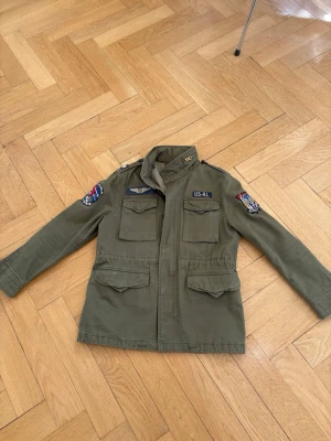 Olivgrön field jacket från Polo Ralph Lauren - Säljer en olivgrön field jacket från Polo Ralph Lauren med schyssta patchar på ärmarna och bröstet. Jackan har fyra stora fickor framtill, axelklaffar och stängs med dragkedja och knappar. Perfekt för dig som gillar militärinspirerad stil. Nypris 2995kr. Säljer den för 2000kr. Storlek 10-12 år.