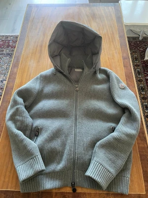 Moncler cardigan  - Säljer en grå stickad cardigan från Moncler med dragkedja framtill och två fickor med dragkedja. Cardigan har en vadderad huva och Moncler-logga på ärmen. Perfekt för dig som gillar exklusiva streetwear-vibbar och vill ha något både snyggt och bekvämt.