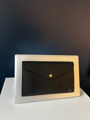 Svart datorväska i ormmönstrat läder - Snygg svart datorväska från Luxie. Man sätter fast den bakom sin dator och kan ha saker i. Finns i beige och svart 