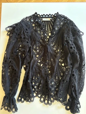 Svart spetsblus från Copenhagen Muse - Supersnygg svart spetsblus från Copenhagen Muse i storlek XS. Blusen har långa ärmar och är gjord i ett genombrutet spetsmaterial med unika cirkeldetaljer och vågig kant. Perfekt för dig som vill sticka ut med en cool och trendig look.