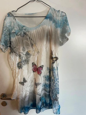 Vit mesh strandtunika med fjärilar - Säljer en luftig vit mesh topp med korta ärmar och blommigt mönster samt färgglada fjärilar. Toppen har blå detaljer upptill och nertill, samt transparent struktur som ger en cool och somrig vibe. Perfekt att styla över ett linne eller bikini. Rök och djurfritt hem. Pruta gärna 🌸