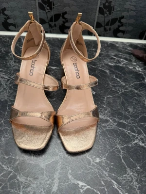 Guldiga klackar från Boohoo - Säljer ett par metallic guldiga pumps från Boohoo med öppen tå, bred klack och remmar över foten samt runt ankeln. Skorna har dragkedja bak för enkel påtagning och en fyrkantig tå. Perfekta för att lyfta vilken outfit som helst.