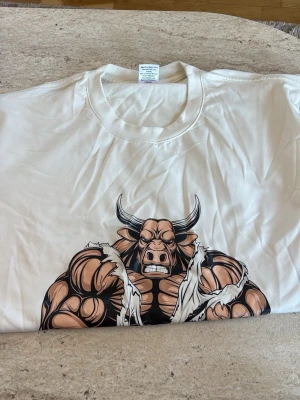 Vit t-shirt med tryck  - Cool vit t-shirt i 100% polyester med ett stort tryck av en tecknad, muskulös och arg tjur på framsidan. Klassisk rund halsringning och korta ärmar. Perfekt för dig som gillar unika och iögonfallande motiv.