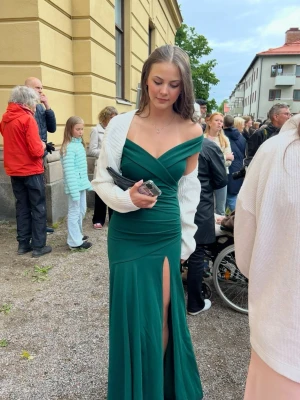 Grön balklänning med slits - Elegant grön långklänning med offshoulder-design och v-ringad urringning. Klänningen har en hög slits framtill och figurnära passform. Perfekt för bal! Endast använd vid ett tillfälle 
