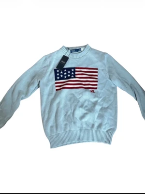 Ljusblå stickad tröja Polo Ralph Lauren - Snygg ljusblå stickad tröja från Polo Ralph Lauren med amerikansk flagga broderad på bröstet och RL-detalj. Tröjan har rund halsringning och ribbstickade muddar vid ärmslut och nederkant. Perfekt för dig som gillar klassisk preppy stil.