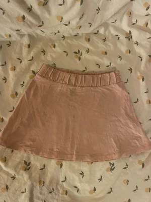 Ljusrosa kjol från Amy & Lucy Barn st 122/128 - Söt ljusrosa kortkjol från Amy & Lucy med resår i midjan. Kjolen har en enkel och stilren design, perfekt för att matcha med olika toppar. Tillverkad i mjukt bomullsmaterial. Barn storlek 122/128