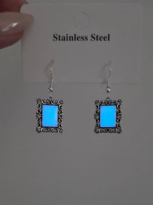 Glow in the dark örhängen vintage ram - Lyser blått! - Unika örhängen i rostfritt stål med rektangulär ram och glow in the dark-effekt. Ramen har detaljerade snirkliga mönster och stenen i mitten lyser blått i mörker och är vit i dagsljus. Perfekt för dig som vill sticka ut med coola accessoarer. Ljus effekten aktiveras av vanligt dagsljusnär du bär dom som vanligt, solljus fungerar också, de lös direkt efter 3 sekunder när jag fotade. Normal användning kan de lysa upp till 12 timmar. Handgjort & tål vatten.