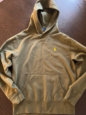 Olivgrön hoodie från Polo Ralph Lauren - Säljer en olivgrön hoodie från Polo Ralph Lauren med klassisk gul broderad logga på bröstet. Hoodien har huva, känguruficka och ribbade muddar vid ärmar och nederkant. Tillverkad i mjuk bomullsblandning, perfekt för en avslappnad stil.