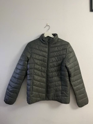 Grå pufferjacka med dragkedja - Snygg grå pufferjacka med hög krage och hel dragkedja framtill. Jackan har ett quiltat mönster med horisontella sömmar och är lätt vadderad för extra värme. Perfekt för kalla vinterdagar och enkel att matcha med olika outfits.