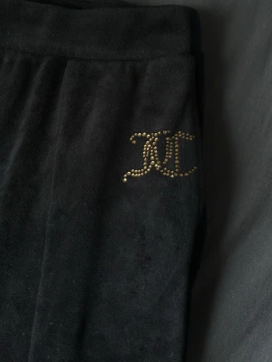 Svarta mjukisbyxor från Juicy Couture - Jag säljer ett par Juicy Couture byxor, svarta med guldiga glitter stenar på ena höften och fickor på sidorna. Obs!! Dem är tyvärr lite trasiga längst ned på benet ( där av priset). Slittet ska vara där på båda benen men de har spruckit upp lite åvanför. Men med sy kunskaper så kan detta gå att laga till ett fint skick. Annars ett bra skick!
