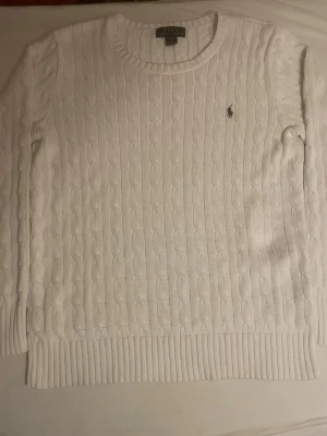 Vit kabelstickad tröja Polo Ralph Lauren - Kabelstickad vit tröja från Polo Ralph Lauren med rund halsringning och diskret broderad logga på bröstet. Tröjan har långa ärmar och ribbade muddar vid ärmslut och nederkant. Klassisk och stilren design i mjukt material. Inga fula fläckar eller defekter 