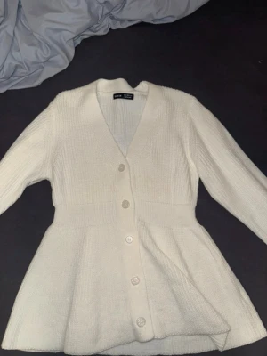 Vit ribbstickad kofta med knappar - Snygg vit ribbstickad kofta med v-ringning och fyra stora knappar framtill. Koftan har en lätt utsvängd passform nedtill och långa ärmar 