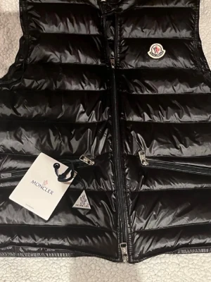 Svart dunväst från Moncler gui - Snygg svart dunväst från Moncler med glansig finish och klassisk Moncler-logga på bröstet. Västen har två dragkedjeförsedda fickor och hög krage. Perfekt för lager-på-lager och streetstyle. Materialet är dunfyllning med yttertyg i nylon. Storlek 2 