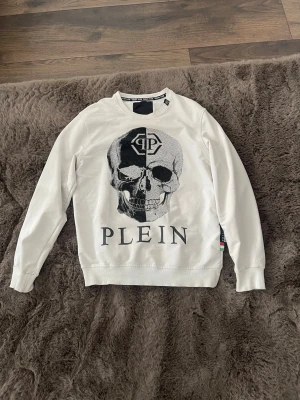 Plilip Plein - Cool vit sweatshirt från Philipp Plein med stor svartvit dödskalle och logga på bröstet. Tröjan har rund hals, långa ärmar och ribbade muddar. Materialet känns mjukt och skönt, perfekt för en streetwear-look.