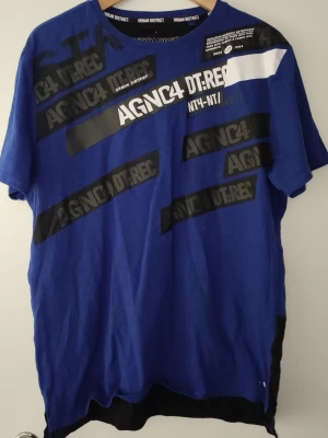 Blå Urban District t-shirt med tryck - Cool blå t-shirt från Urban District med svart och vitt grafiskt tryck framtill. T-shirten har korta ärmar och svart panel nertill på baksidan. Perfekt för dig som gillar streetwear och vill sticka ut lite extra.