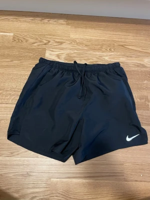Svarta träningsshorts från Nike - Säljer ett par svarta träningsshorts från Nike med elastisk midja och snörning. Shortsen har en klassisk loose passform och är tillverkade i ett lätt, syntetiskt material. Perfekta för gymmet eller löprundan. Nike-logga på vänster ben.