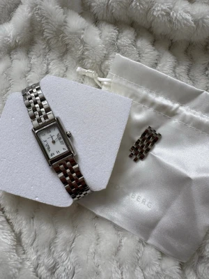 Silverfärgad klocka från Mockberg - Den trendiga, stilrena klocka i rektangulär form från Mockberg i silverfärg med länkarmband i metall. Urtavlan är vit med romerska siffror och tunna svarta visare. Kommer med extra länkbitar och en vit tygpåse med Mockberg-logga. Nypris 1900kr