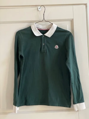 Grön långärmad Moncler piké - Mörkgrön långärmad pikétröja från Moncler med vit krage och vita muddar. Klassisk knappslå framtill och Moncler-logga på bröstet. Tillverkad i mjuk bomull för en bekväm känsla och stilren look.