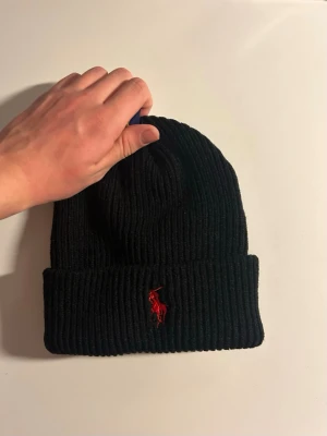 Svart ribbstickad mössa från Polo Ralph Lauren - Säljer en svart ribbstickad mössa från Polo Ralph Lauren med uppvikt kant och den klassiska röda logotypen broderad framtill. Mössan är i mjukt material och har en enkel, stilren design som passar till det mesta. Helt oanvänd. No size