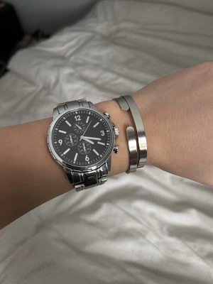 Silverfärgad klocka från Pier One - Stilren klocka från Pier One med svart urtavla och silverfärgat armband i metall. Klockan har kronografdetaljer och tydliga vita siffror samt markeringar. Perfekt accessoar för dig som gillar en klassisk och modern look.