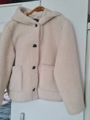 Beige teddyjacka med huva från Zara - Mysig beige teddyjacka från Zara med huva och stora framfickor. Jackan har svarta knappar framtill och är tillverkad i ett fluffigt, mjukt material som ger en avslappnad och trendig look. Perfekt för kyliga dagar när du vill vara både varm och snygg.