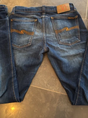 Blå jeans från Nudie Jeans - Säljer ett par klassiska blå jeans från Nudie Jeans med snygga kontrastsömmar och ikonisk brodyr på bakfickorna. Jeansen har normal passform och är tillverkade i slitstarkt denimtyg. Perfekta för en avslappnad och trendig look. L32 W29