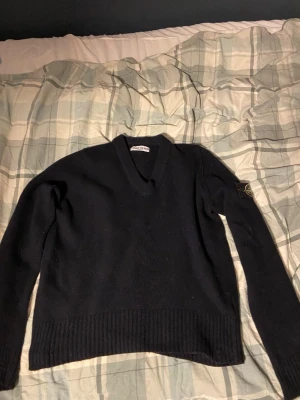 Svart stickad tröja från Stone Island - Svart stickad tröja från Stone Island med V-ringning och ribbstickade muddar. Klassisk logotyp-patch på vänster ärm. Perfekt för dig som gillar stilrena och tidlösa plagg med streetwear-vibe.