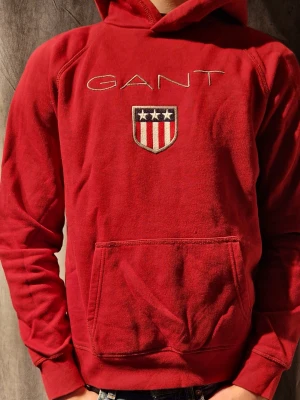 Röd hoodie från GANT - Snygg röd hoodie från GANT med broderad logga. Klassisk känguruficka framtill och huva. Mjuk bomullskvalitet och ribbade muddar vid ärmslut och nederkant.