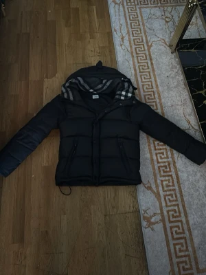 Svart pufferjacka med avtagbara ärmar - Säljer en svart pufferjacka från Burberry i storlek S med avtagbara ärmar så den kan användas som väst. Jackan har huva med klassiskt rutigt foder, två dragkedjefickor framtill och en puffig siluett. Perfekt för dig som vill ha en flexibel och snygg jacka med streetkänsla. Jag säljer denna jacka därför att den var för liten på mig😎