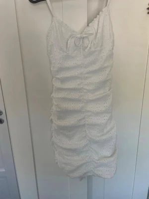 Vit kort klänning med broderi - Söt vit kortklänning med smala axelband och broderade detaljer. Klänningen har en rosett framtill, omlottdesign vid bysten och rynkade partier längs hela längden för en snygg passform. Perfekt för varma dagar och sommarens alla tillfällen.