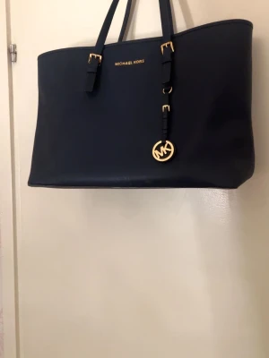 Svart Michael Kors handväska - Stilren svart handväska från Michael Kors i slätt läder med två axelband och guldfärgade detaljer. Väskan har ett klassiskt MK-hänge och logga framtill. Perfekt för dig som gillar en enkel men lyxig look. Väskan är ganska så oanvänd, däremot finns en fläck i väskan men som inte syns när man har på sig den. 