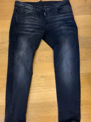 Svarta skinny jeans från Dsquared2 - Säljer ett par svarta skinny jeans från Dsquared2 med slitningar och diskreta färgstänk. Jeansen har klassisk femficksdesign, låg midja och stretchigt material för en bekväm passform. Perfekta för en trendig streetstyle-look.