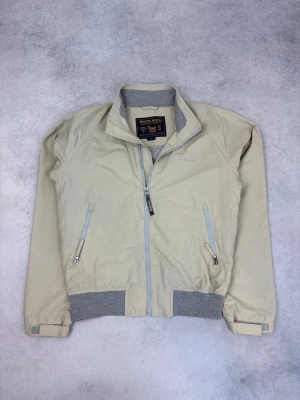 Beige vindjacka från Woolrich - Stilren beige vindjacka från Woolrich med grå ribbade muddar och krage. Jackan har två snedställda fickor med dragkedja och diskret logga på bröstet. Perfekt för blåsiga dagar och enkel att matcha med olika outfits.