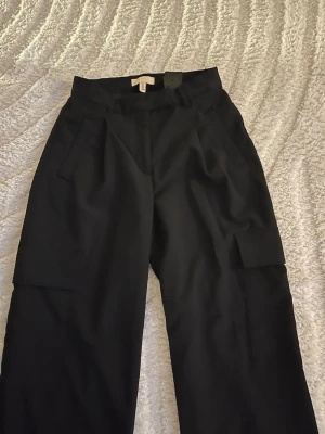 Svarta cargobyxor från H&M - Stilrena svarta cargobyxor från H&M med raka ben och stora sidofickor. Byxorna har hög midja och praktiska fickor framtill. Perfekta för en avslappnad och trendig look. Materialet känns mjukt och har en lätt stretch för extra komfort.