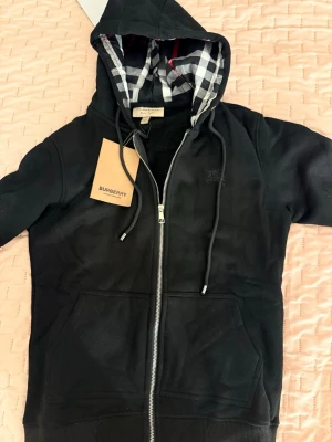 Svart Burberry Zipper - Snygg svart hoodie från Burberry med dragkedja framtill och klassiskt rutigt foder i huvan. Broderad Burberry-logga på bröstet och två stora fickor framtill. Tillverkad i mjukt bomullsmaterial och har justerbara snören i huvan.