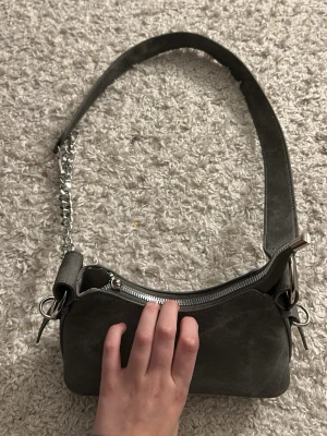 Grå axelväska  - Hej, jag säljer min gråa axelväska eftersom den inte kommer till användning. Köpt på SHEIN men ändå en väska som är med i trenderna. Den har en ”metall kedja” som man kan spänna in och ut. Dragkedjan, asså den man drar dragkedjan med, har tappat den delen som ni ser på sista bilden. Men de går ändå och öppna o stänga utan problem.