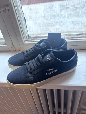 Svarta Saint Laurent sneakers - Snygga svarta sneakers från Saint Laurent med vit logga på sidan och vita sulor. Skorna har klassisk låg profil, svarta skosnören och är tillverkade i canvas och läder. Perfekta för dig som gillar stilrena och exklusiva sneakers.