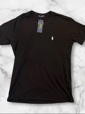 Svart t-shirt från Polo Ralph Lauren - Klassisk svart t-shirt från Polo Ralph Lauren med den ikoniska vita logobrodyr på bröstet. T-shirten har rund halsringning och korta ärmar. Tillverkad i mjuk bomull för en skön och avslappnad look.