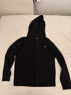 Svart hoodie från Polo Ralph Lauren - Svart hoodie med dragkedja från Polo Ralph Lauren. Klassisk design med vit broderad logga på bröstet, två fickor fram och justerbar huva med snören. Mjuk bomullsblandning som är skön att ha på sig. Perfekt för en chill och stilren look.