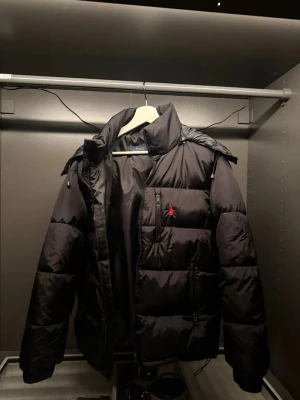 Svart pufferjacka från Polo Ralph Lauren - Säljer en svart pufferjacka från Polo Ralph Lauren med röd broderad logga på bröstet. Jackan har hög krage, dragkedja framtill och flera fickor. Tveka inte att höra av dig vid frågor och priset går att diskutera vid snabb affär😁