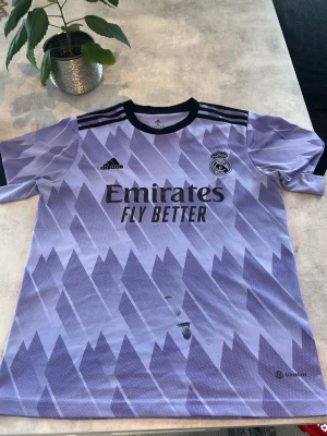 Real Madrid matchtröja lila Adidas - Snygg Real Madrid matchtröja i lila med grafiskt mönster och svarta detaljer. Adidas-logga och klubbmärke på bröstet samt 'Emirates Fly Better' tryckt framtill. Tillverkad i lätt och ventilerande material, perfekt för fotboll eller träning.