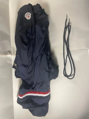 Marinblå vindjacka från Moncler - Snygg marinblå vindjacka från Moncler med huva och dragkedja. Jackan har Moncler-logga på bröstet och en randig detalj i rött, vitt och grått vid nederkanten. Tillverkad i lätt, glansigt material som står emot vind. Perfekt för dig som vill ha en sportig och stilren look.