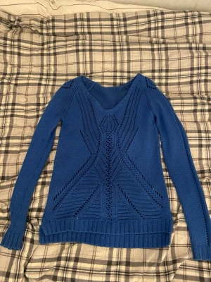 Blå stickad tröja med mönster - Snygg blå stickad tröja med V-ringning och detaljerat hålmönster framtill. Tröjan har långa ärmar och ribbade muddar vid ärmslut och nederkant. Perfekt för dig som gillar en unik och trendig look. Materialet känns mjukt och bekvämt. Labb är bortklippt.