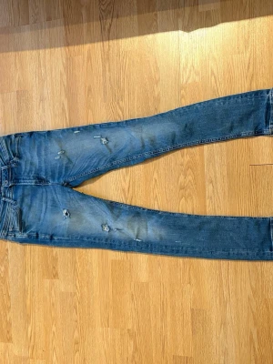 Blå slitna jeans - Säljer ett par blåa jeans med slitningar och distressed detaljer. Jeansen har klassisk femficksmodell och är i stretchigt denimtyg. Perfekta för en avslappnad och trendig look.