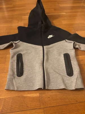 Nike hoodie i svart och grå - Snygg hoodie från Nike i svart och ljusgrått med dragkedja framtill och två fickor med svarta dragkedjor. Klassisk Nike-logga på bröstet och huva för extra stilpoäng. Perfekt för dig som gillar sportig och avslappnad look.