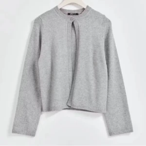 Ljusgrå kofta med knapp - Stilren ljusgrå kofta med rund halsringning och en knapp upptill. Koftan har långa ärmar och ribbade kanter vid ärmslut och nederkant. Perfekt att slänga över en topp för en enkel och clean look.