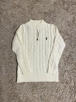 Vit kabelstickad tröja Polo Ralph Lauren - Kabelstickad vit tröja från Polo Ralph Lauren med halv dragkedja och ribbade muddar. Tröjan har det klassiska mörkblå logobroderiet på bröstet och är tillverkad i mjukt material. Perfekt för lager på lager och en clean look.